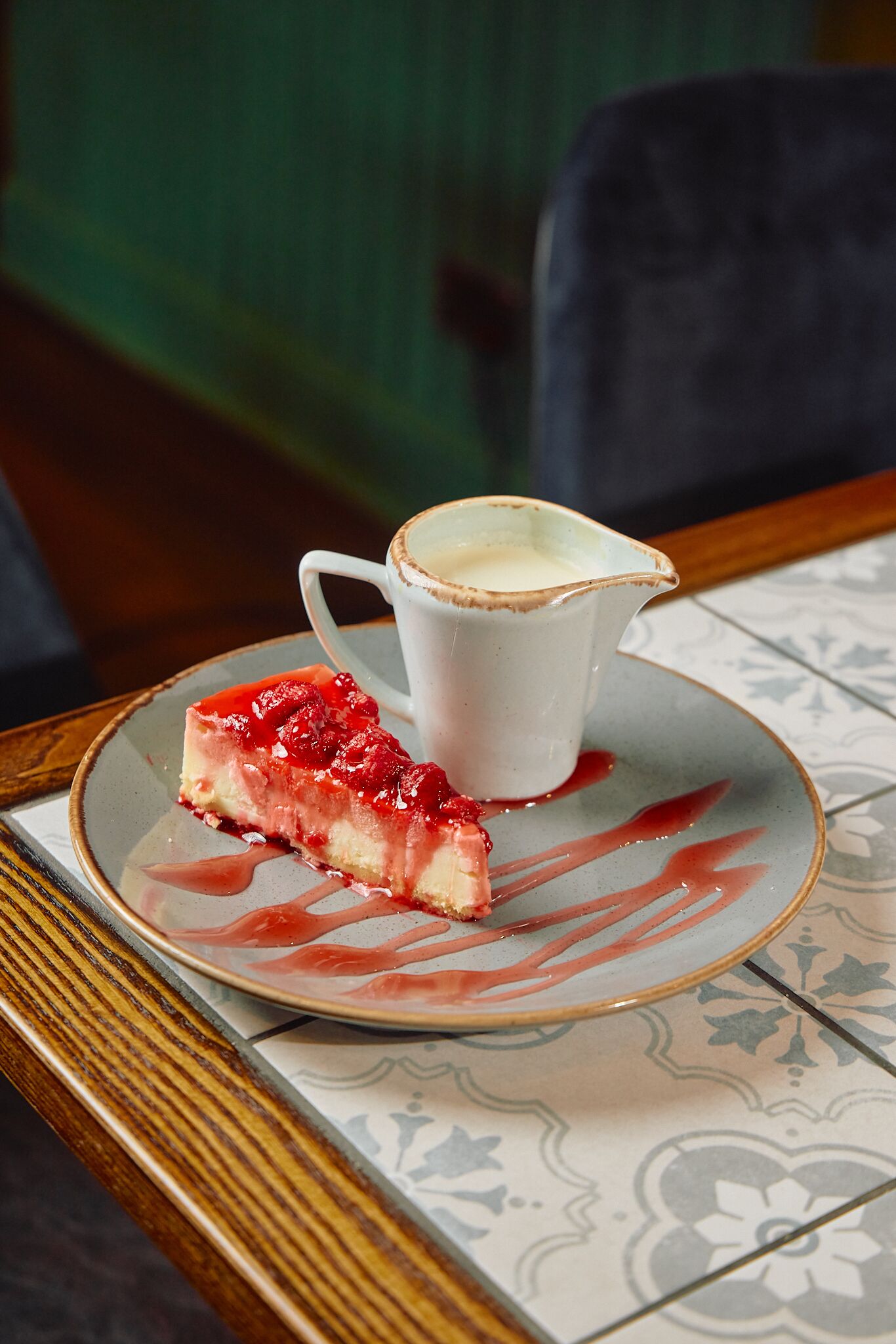 A dessert plate with a slice of cheesecake and a small pitcher of cream on a patterned tablecloth.