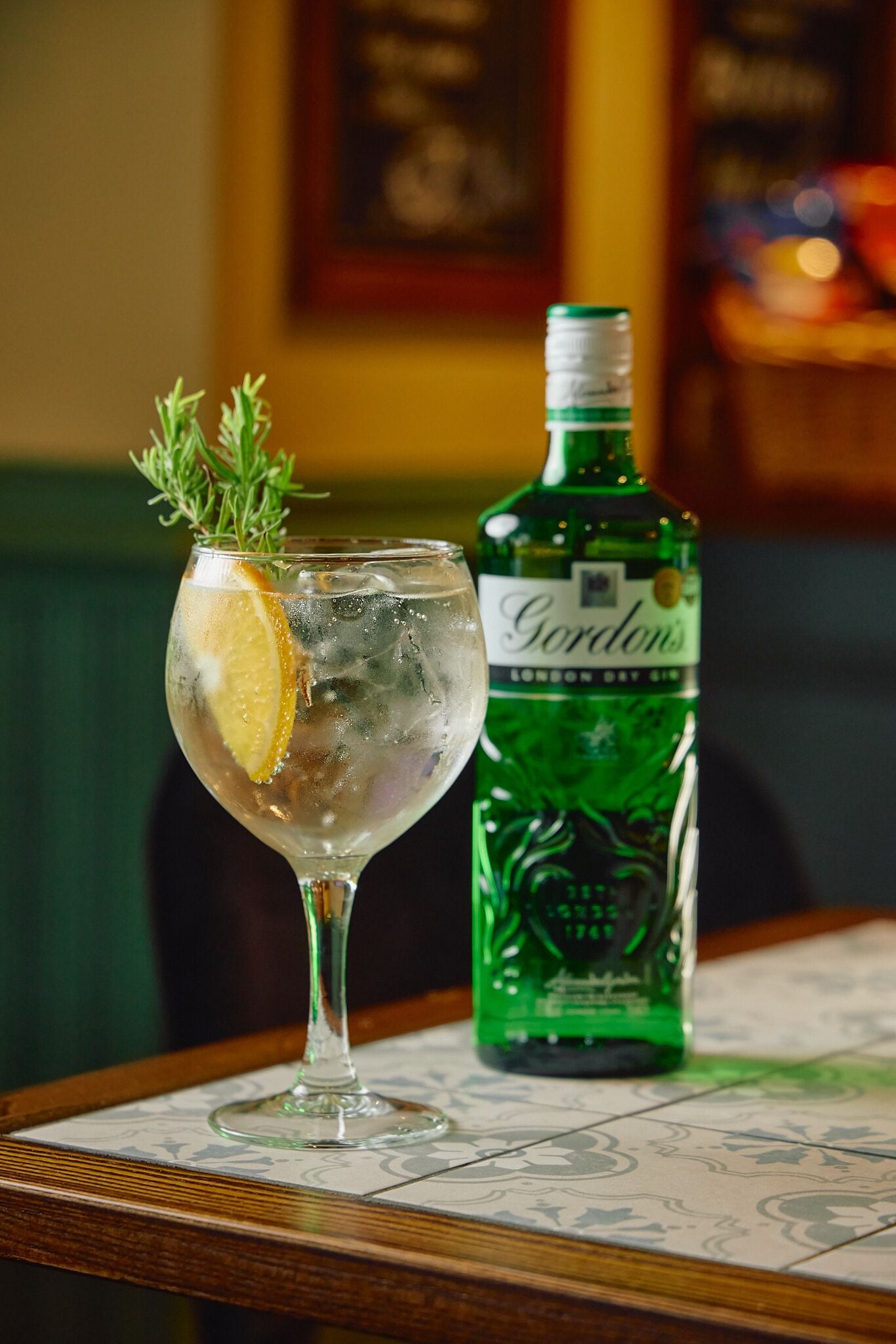 A cocktail glass with a lemon slice and a sprig of rosemary, next to a bottle of Gordon's London Dry Gin on a table with a patterned tablecloth.