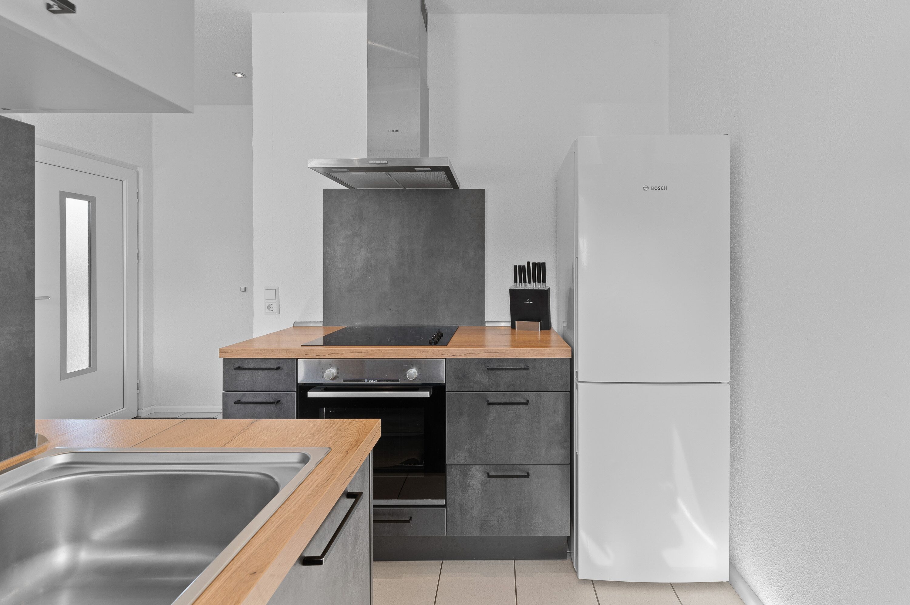 A modern kitchen with a stainless steel sink, a built-in oven, a white Bosch refrigerator, and a wooden countertop.