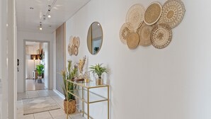 A hallway with a white tiled floor, a glass console table with plants, a mirror, and woven wall art. - LUMIFLATS (Mahlberg)