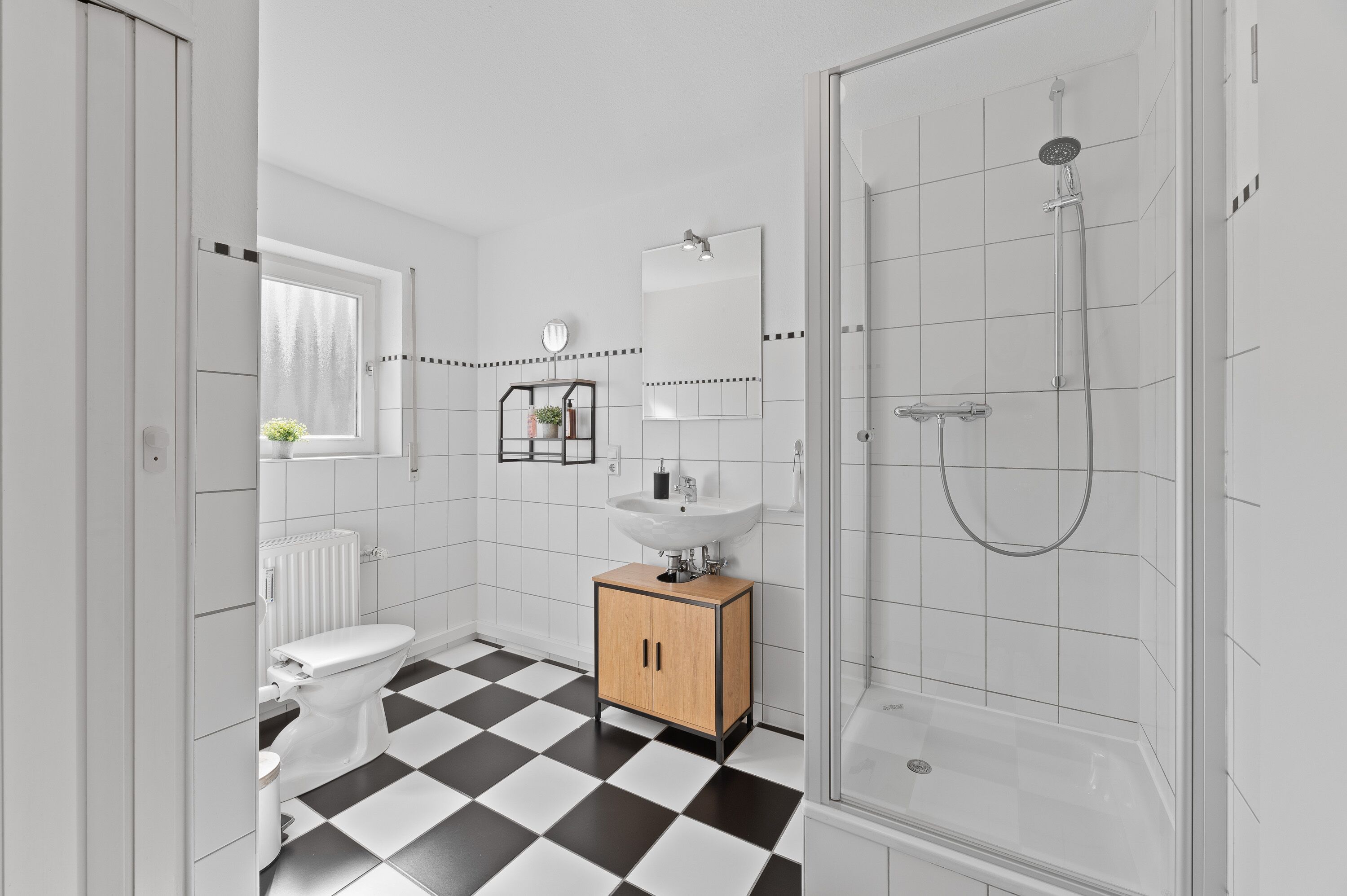 Une salle de bain moderne avec un sol à carreaux noirs et blancs, des toilettes blanches, un meuble en bois, un lavabo, un miroir et une douche.