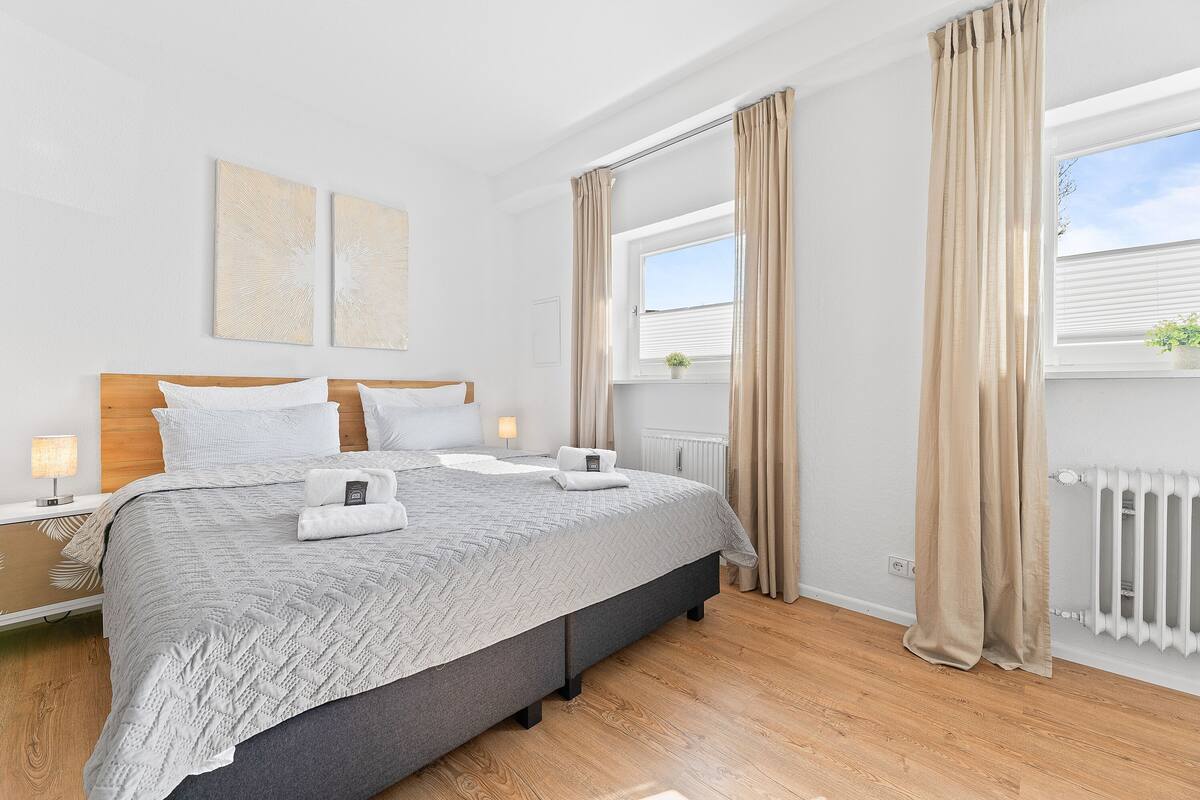 Un dormitorio moderno con una cama grande, lámparas de noche, una ventana con cortinas y un radiador.