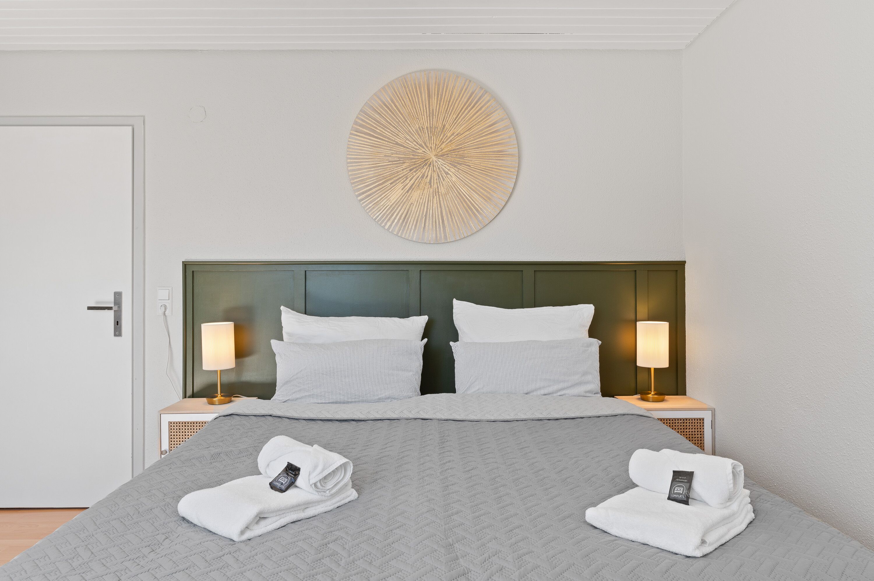 A neatly made bed with a grey quilt, two white pillows, and two bedside lamps.