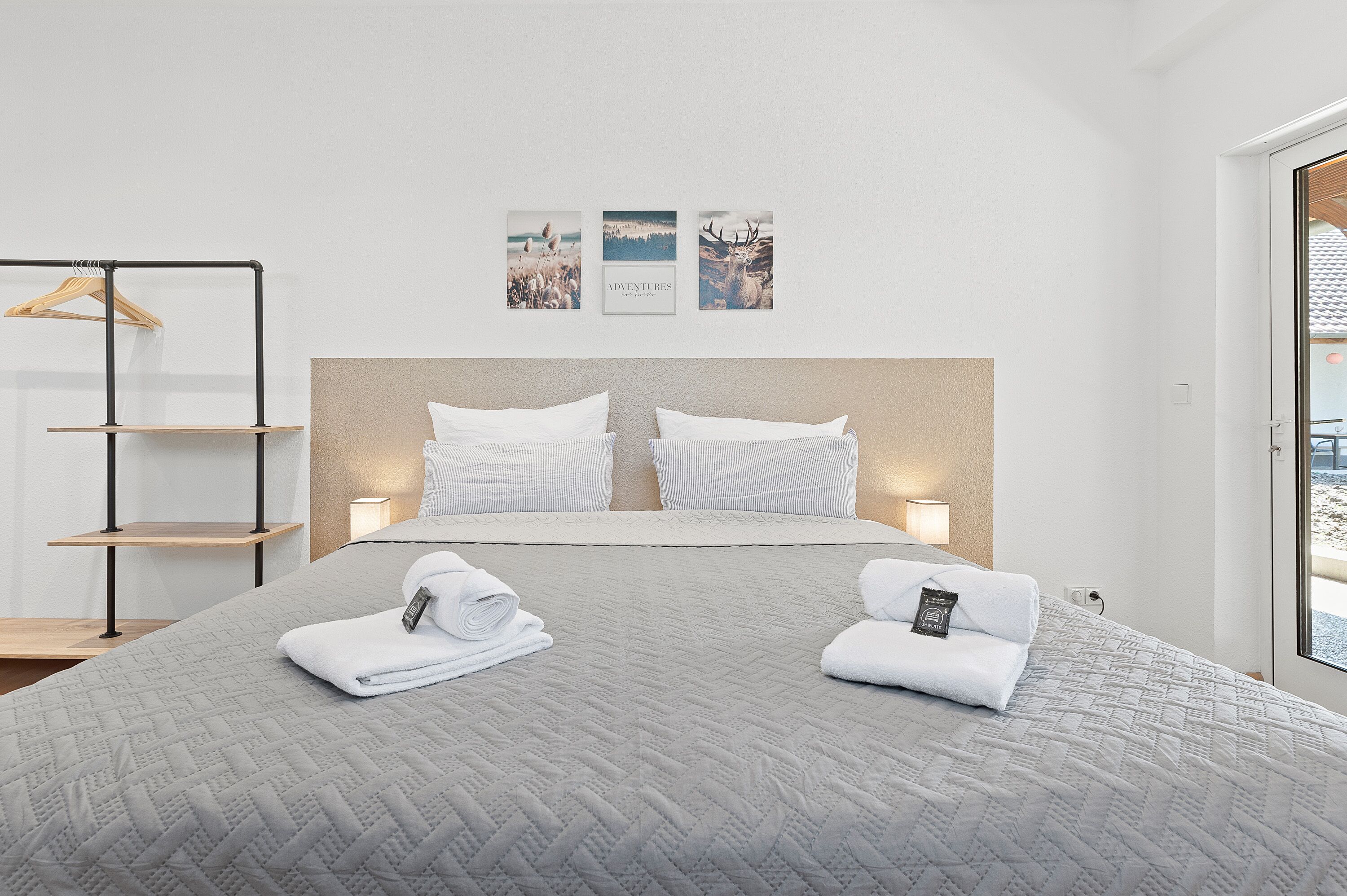 A neatly made bed with a grey quilt, two white pillows, and rolled white towels on the bed. There is a wooden shelf with a hanging garment rack to the left. Three framed pictures are hung on the wall above the bed.