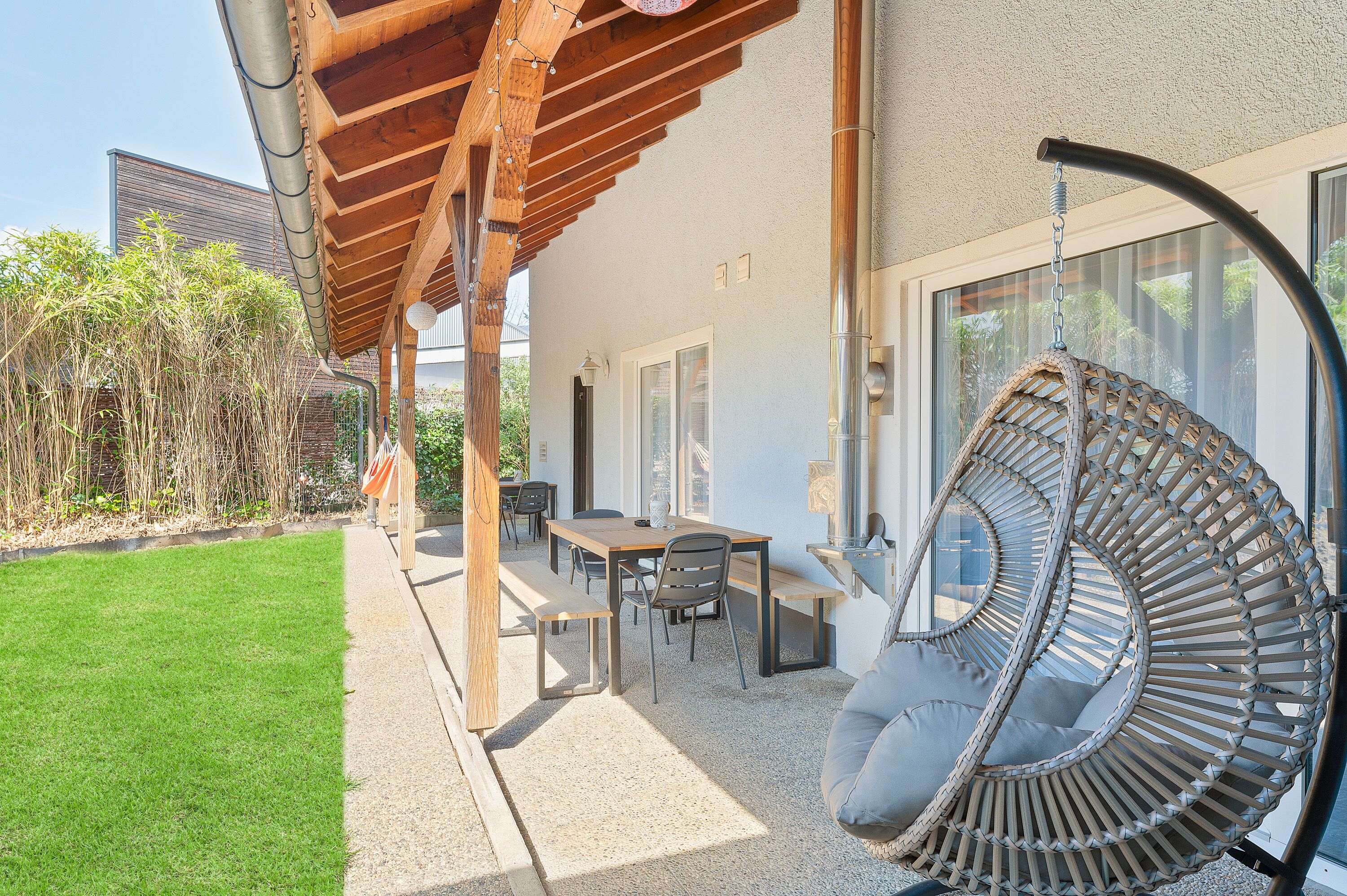 Une terrasse couverte avec une pergola en bois, un fauteuil suspendu en rotin, une table avec des chaises et, au loin, une vue sur un bâtiment.