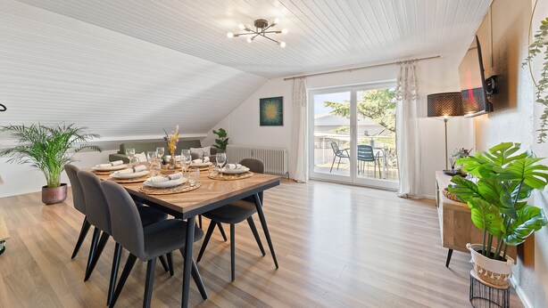 A dining area with a wooden table set for four, surrounded by chairs, a ceiling light fixture, and a view of the outdoors through a sliding glass door. - LUMIFLATS (Mahlberg)