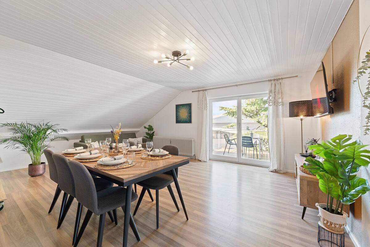Un comedor con una mesa de madera dispuesta para cuatro personas, rodeada de sillas, un lámpara en el techo y vistas al exterior a través de un ventanal corredera.