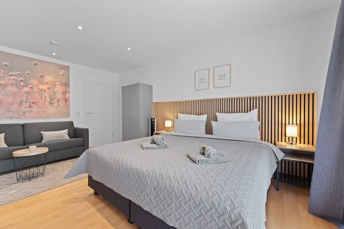 Habitación de hotel moderna con una cama grande, un sofá gris, una mesita pequeña y una obra de arte en la pared.