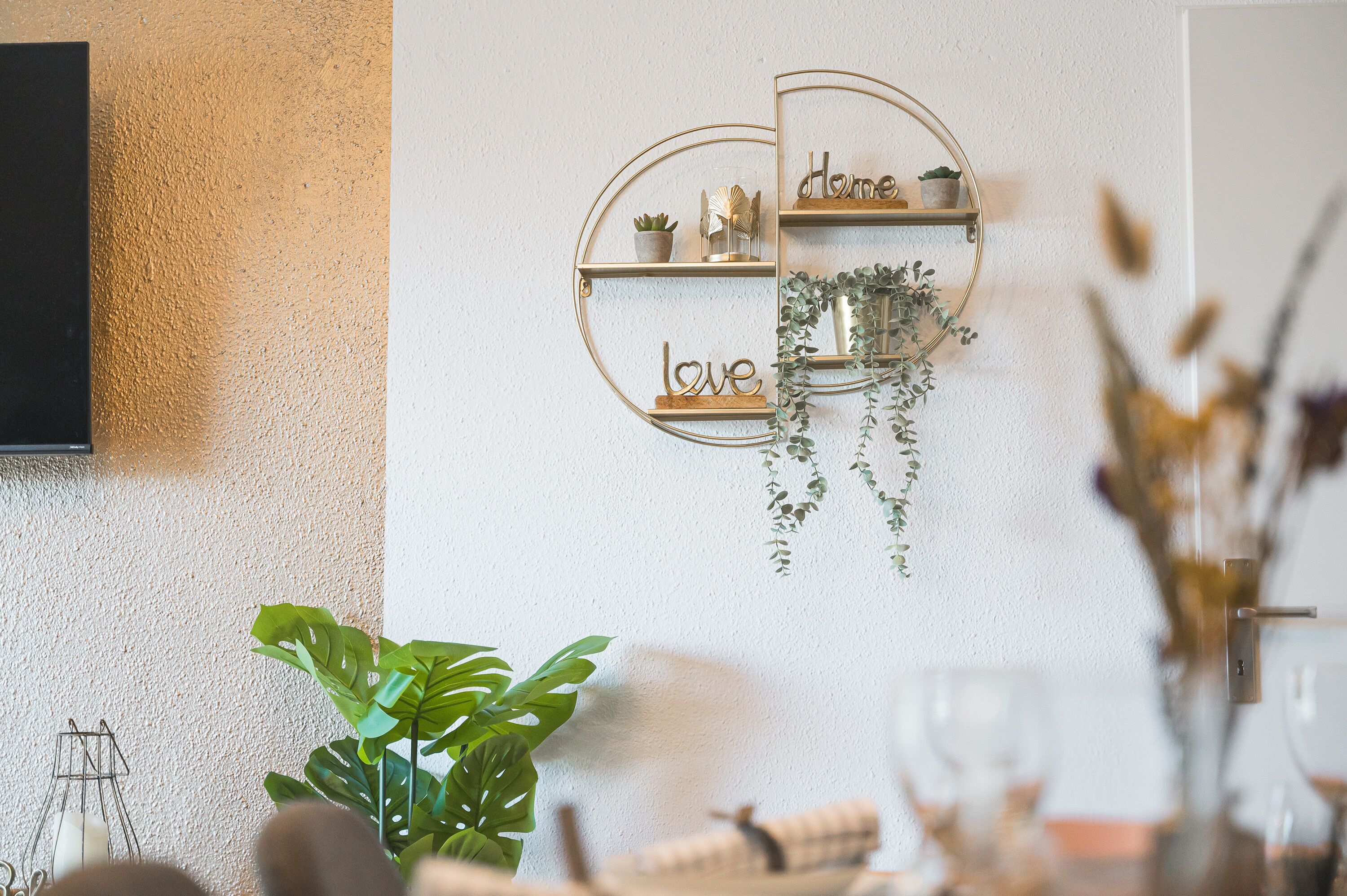 Eine an der Wand befestigte, runde Ablagefläche mit den Aufschriften "home", "love" und "dine", zusammen mit kleinen Topfpflanzen und Dekorationselementen.