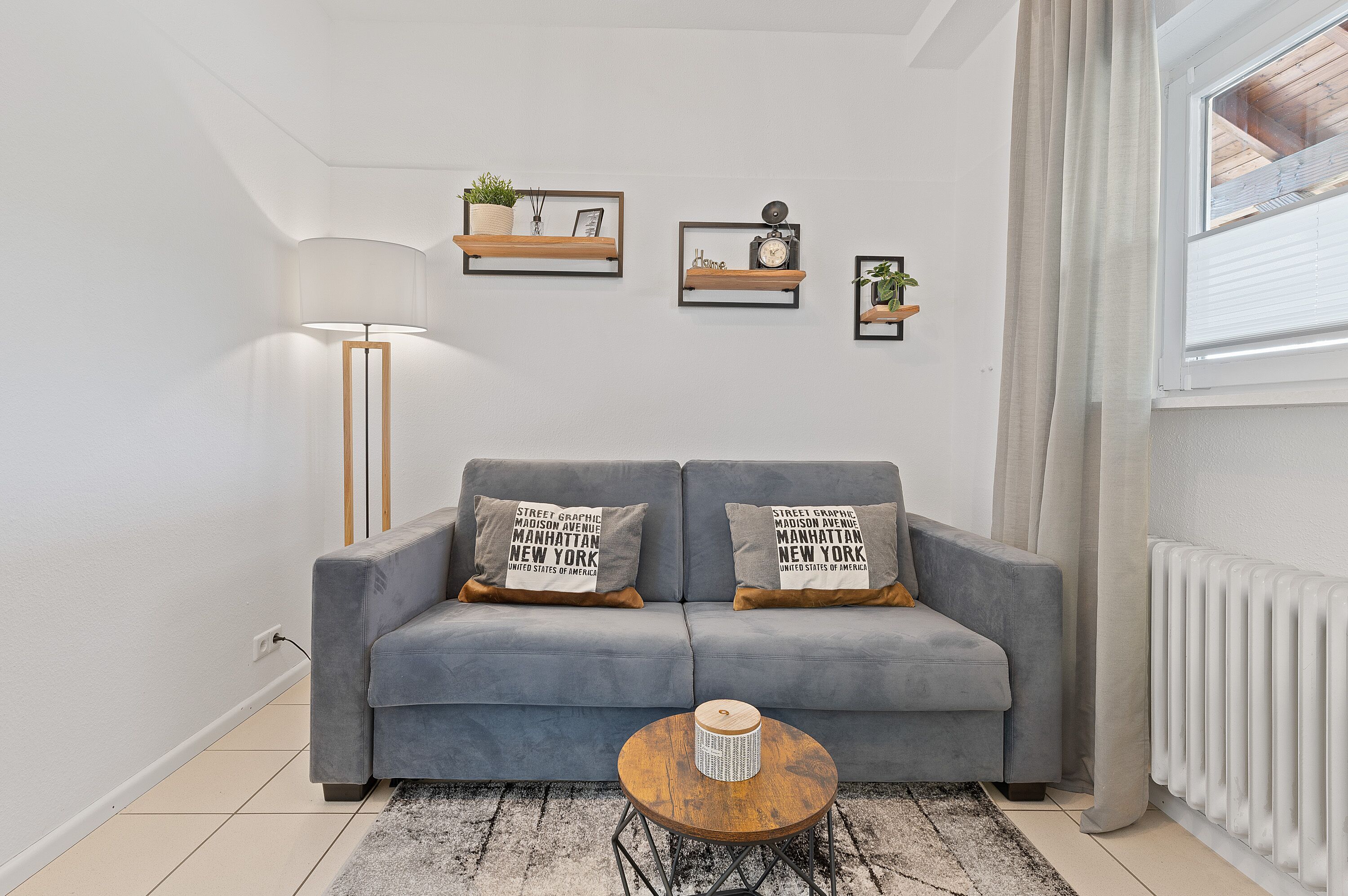 A modern living room with a grey sofa, two decorative pillows, a round coffee table, and three wall-mounted shelves with various items.