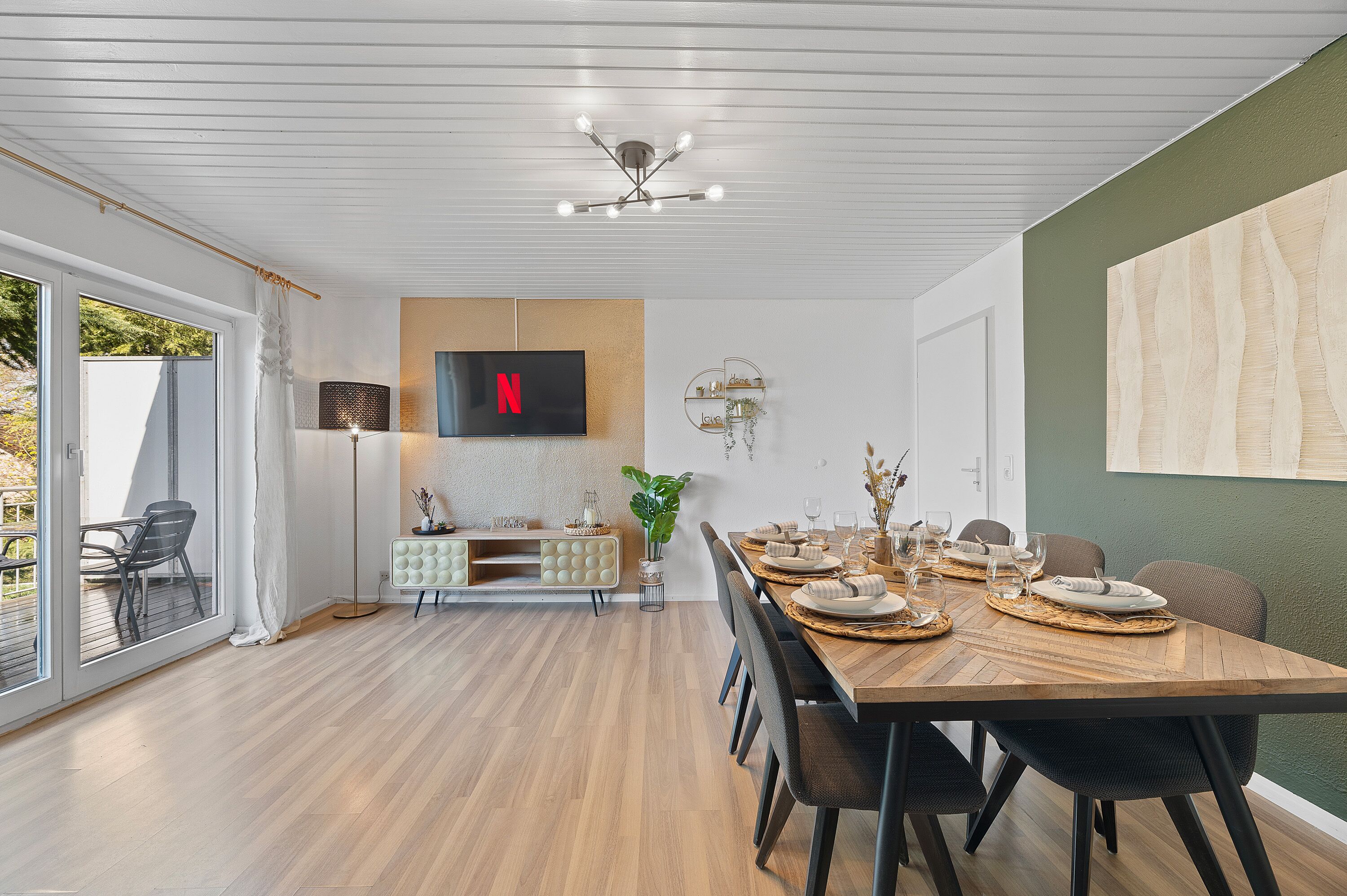 Un comedor con una mesa de madera dispuesta para seis personas, un televisor montado en la pared y un ventanal con cortinas.