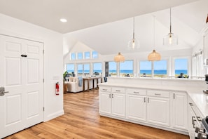 A spacious kitchen with white cabinetry, a wooden floor, and large windows offering a view of the beach.