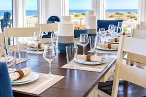 A dining table set for a meal with clear glassware, plates, and napkins, overlooking a beach view.