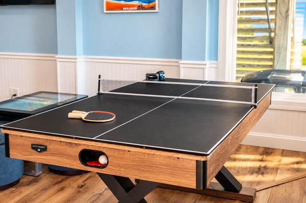 A ping pong table with a paddle, a ball, and a table tennis net in a room with blue walls and a window.