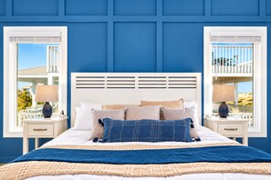A bedroom with a white headboard, two bedside tables, a bed with multiple pillows, and two windows with white frames and blinds.