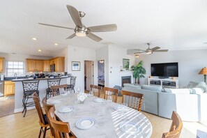 Un vaste espace de vie comprenant un coin repas avec une table pour six personnes, une cuisine avec des meubles en bois, un téléviseur et un ventilateur de plafond.
