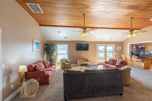 A spacious living room with two sofas, a coffee table, and a TV. There are ceiling fans, a large window, and a view of the outdoors.