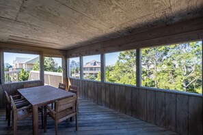 A covered outdoor area with a wooden table and chairs, large windows, and a view of residential buildings and trees.