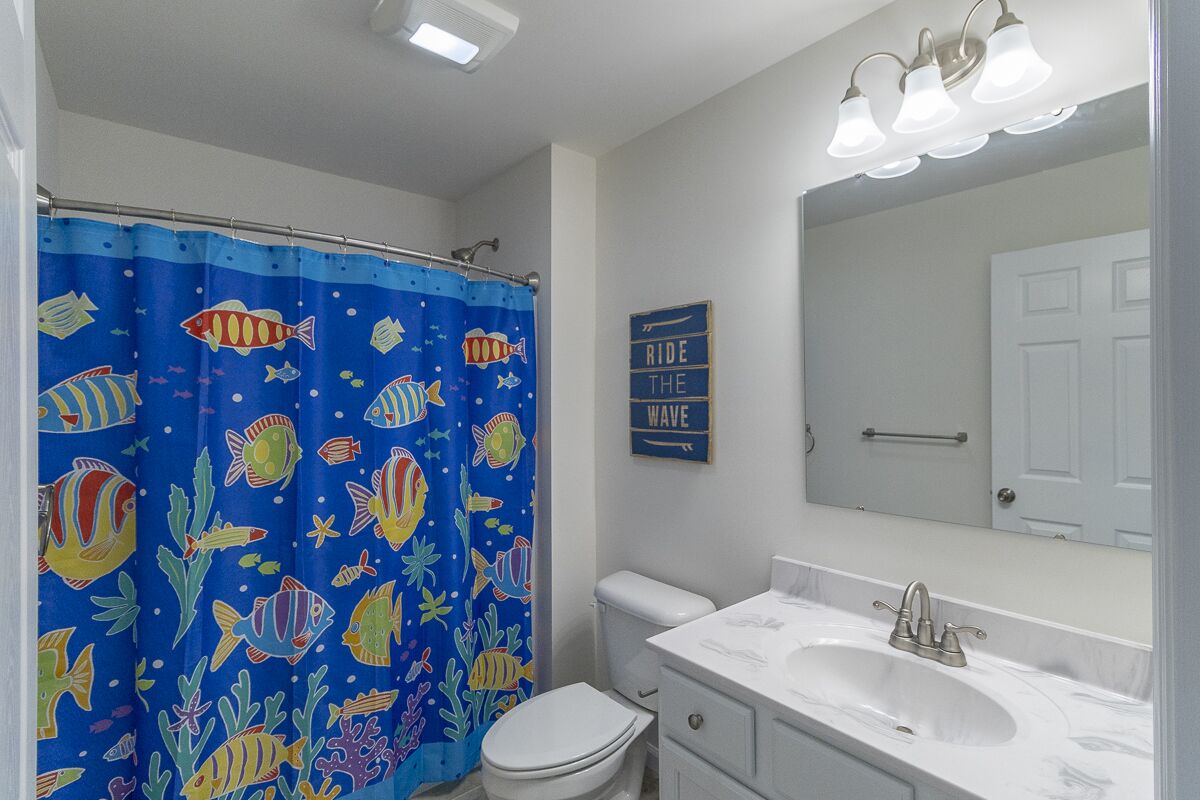 Une salle de bain avec un rideau de douche bleu orné de motifs de poissons, des toilettes blanches, un lavabo avec un miroir au-dessus et un panneau indiquant : « SURFEZ SUR LA VAGUE ».