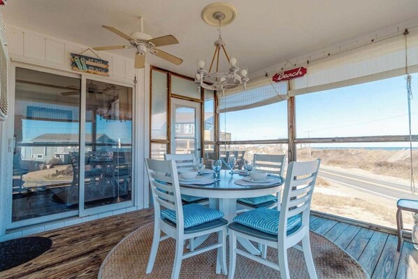A dining area with a round table and chairs, a ceiling fan, and a view of the beach.
