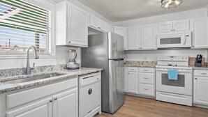 A modern kitchen with white cabinetry, stainless steel appliances, a granite countertop, and a window with blinds. - Throw Em Back Inn, Dog Friendly! (Port Aransas)