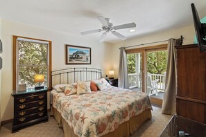 Une chambre à coucher comprenant un lit, un ventilateur de plafond, une fenêtre avec des stores, une table de chevet, une commode et un balcon avec vue sur les arbres.
