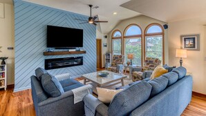 A spacious living room with a large flat-screen TV mounted on a blue wall, a ceiling fan, and a large window with arched top. - Spacious Oceanside Home w/ Pool, Hot Tub, Tiki Bar, & Rec Room (Corolla)
