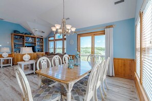 Un espace repas avec une table en bois et des chaises blanches, un lustre et une vue dégagée grâce à de grandes fenêtres.