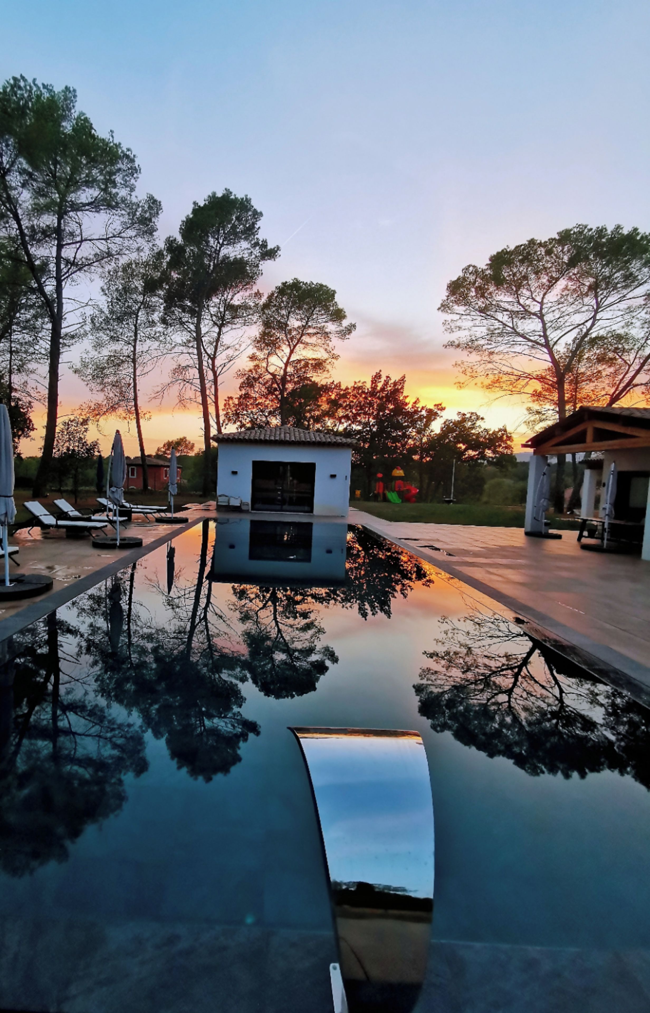 A swimming pool with lounge chairs and umbrellas, surrounded by trees and a small building.