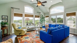 A spacious living room with a blue sofa, a beige armchair, and a coffee table. There are large windows with blinds, a ceiling fan, and a view of greenery outside.