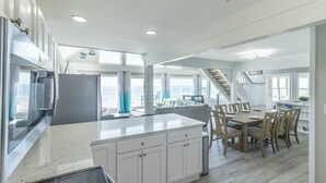 Una cocina amplia con una isla grande, un comedor con sillas de madera y vistas al océano a través de ventanas amplias.