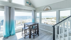 Una habitación con una mesa y sillas de madera, ventanas amplias con vista a la playa y una barandilla blanca.