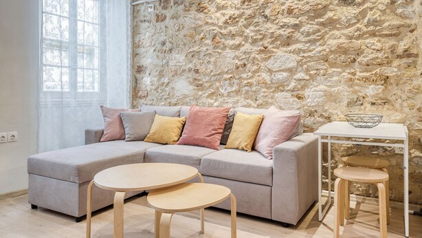 A modern living room with a stone wall, a grey sectional sofa with cushions, a small white table with a bowl, and a window with white curtains. - The Monastery apartment (Corfu)