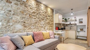 A modern living area with a stone wall, a sofa with cushions, a wooden table, and a kitchen in the background. - The Monastery apartment (Corfu)