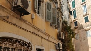 A narrow street with buildings featuring traditional architecture, including shutters and air conditioning units. - The Monastery apartment (Corfu)