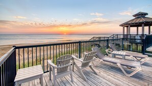 Une terrasse en bois avec des chaises longues et une table, donnant sur une plage au coucher du soleil.