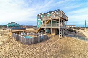 Uma casa de praia de dois andares com um deck de madeira, piscina e um espaço de estar coberto.