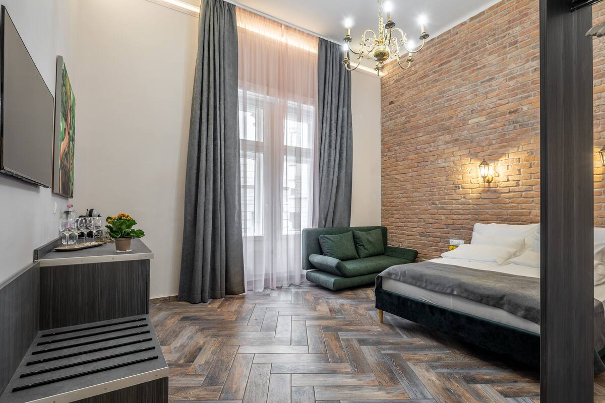 A modern bedroom with a herringbone wood floor, a green sofa, a bed with white linens, a nightstand with glasses and a plant, and a brick accent wall.
