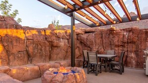 Un patio exterior con una chimenea, mesa y sillas de madera, y una pérgola con luces colgantes.