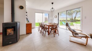 Ein modernes Wohnzimmer mit Kamin, einem Esstisch mit Stühlen, einem Schaukelstuhl und großen Fenstern mit Blick ins Freie.