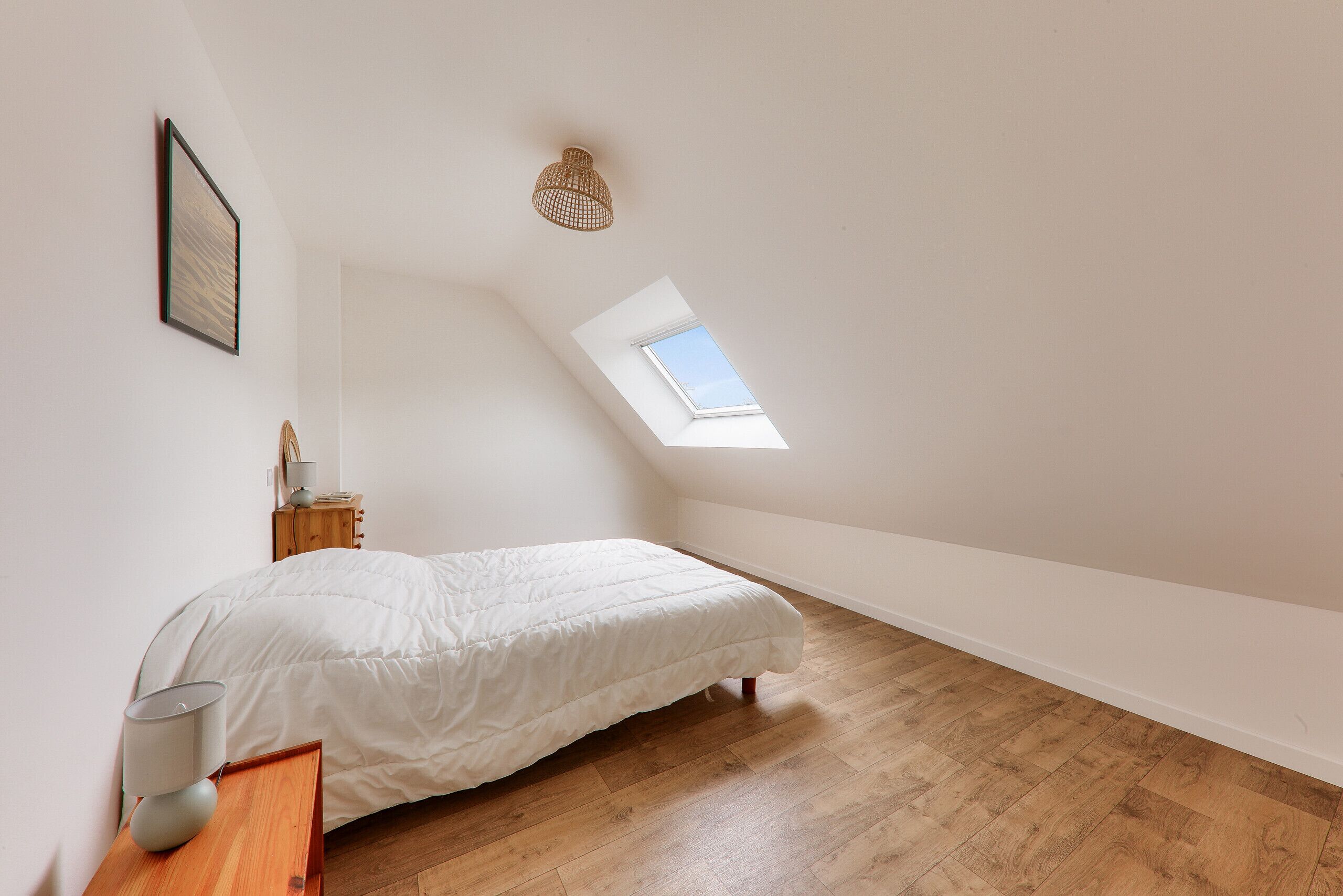 A bedroom with a sloped ceiling, a skylight, a bed, a bedside table with a lamp, and a small wooden nightstand.