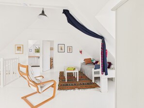 Een kamer met een wit ledikant, een houten schommelstoel, een bed met een kleurrijk dekbed en een klein tafeltje met een groen boek.