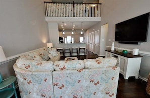 A living room with a sectional sofa, a dining area with bar stools, a flat-screen TV, and a staircase leading to an upper level.