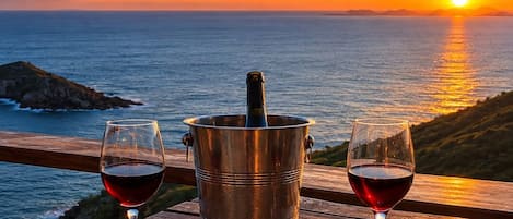 A sunset view with a wooden table featuring two glasses of red wine and a metal bucket with a bottle.