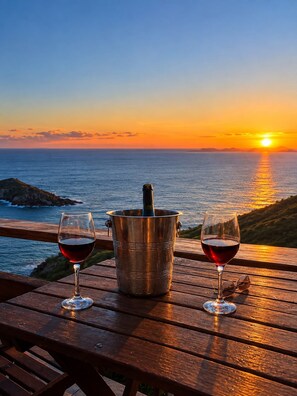 A sunset view with a wooden table featuring two glasses of red wine and a metal bucket with a bottle. - Pousada Sentinelas do Mar (Arraial do Cabo)