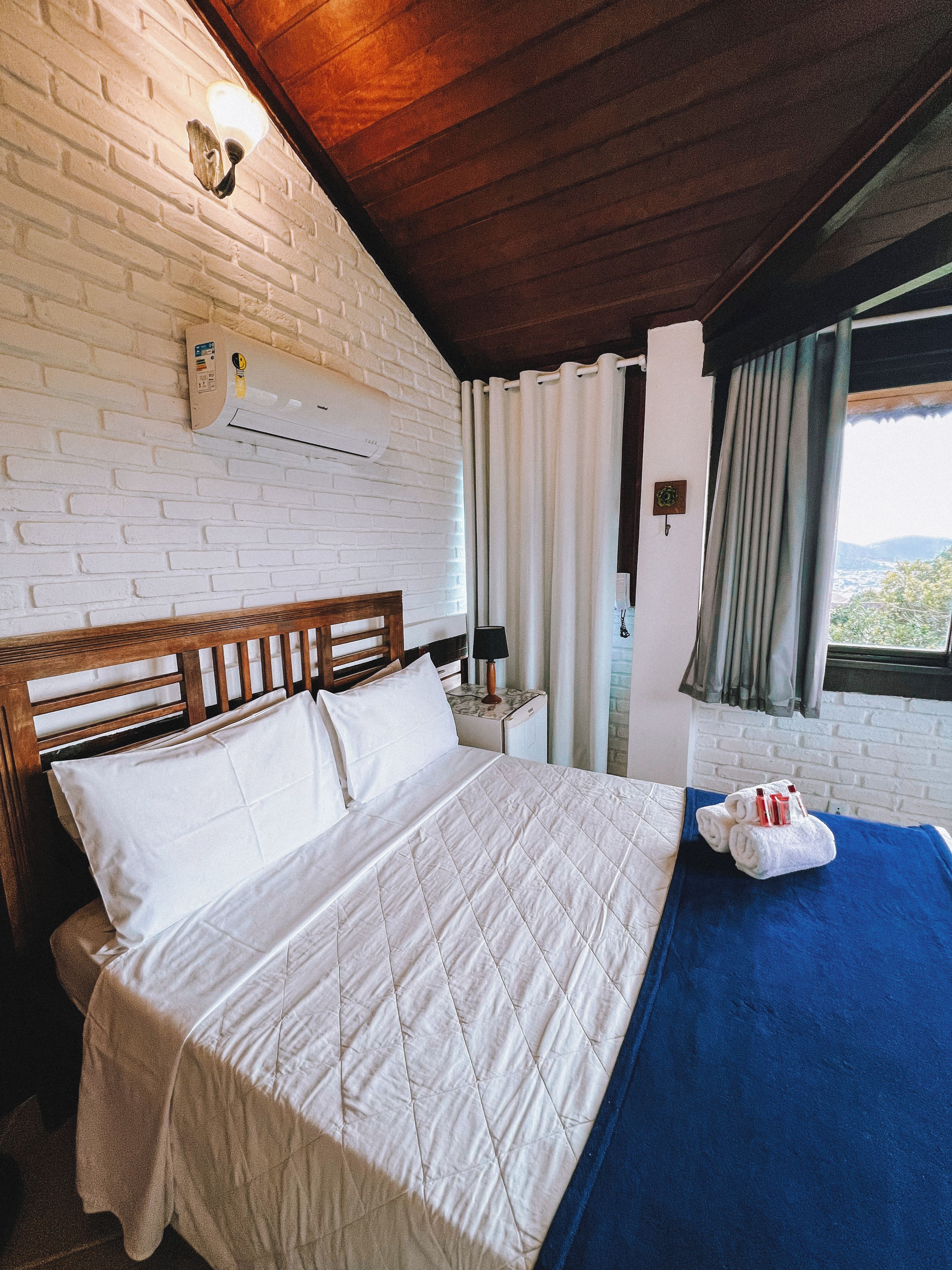 Habitación de hotel con cabecera de madera, colcha blanca acolchada, colcha azul decorativa, pared blanca con diseño de ladrillo y una ventana con cortinas.
