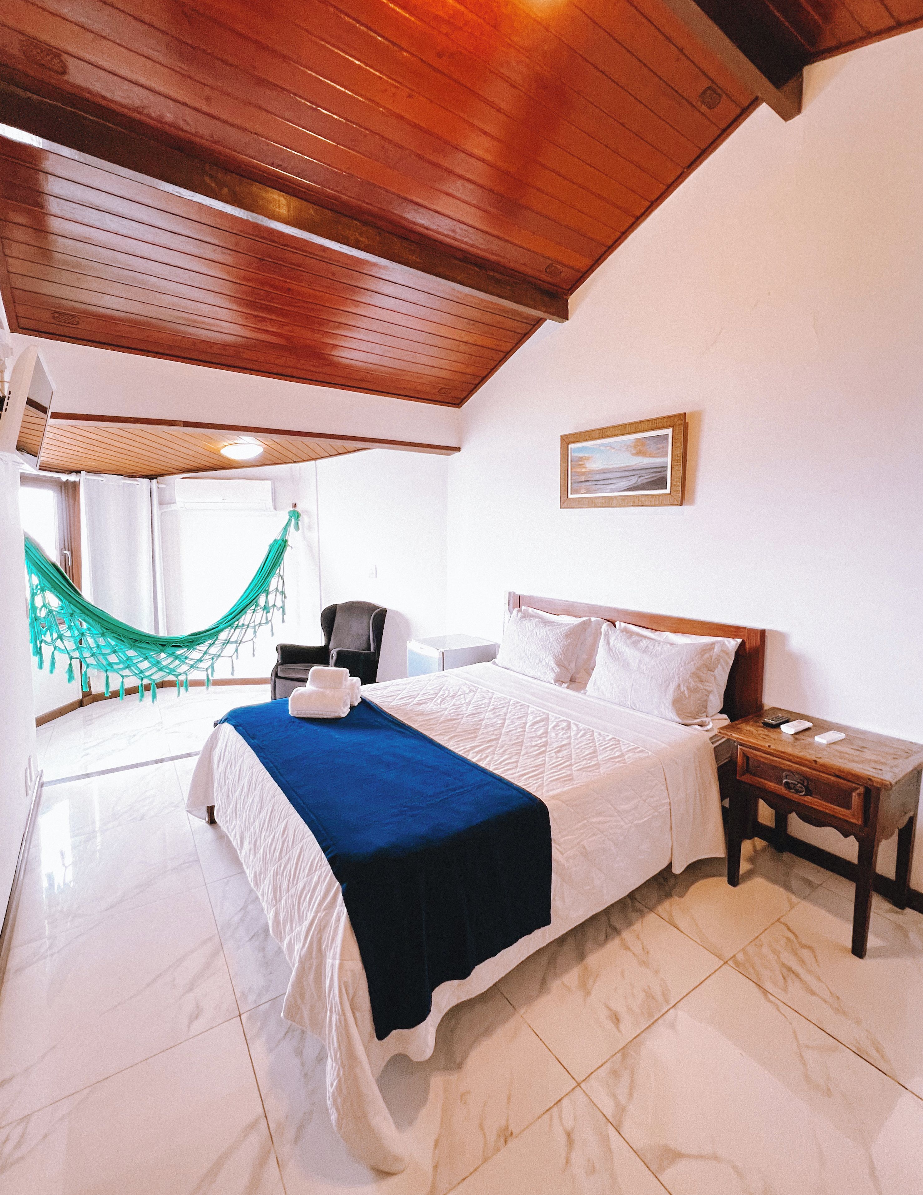 A hotel room with a bed, a nightstand, a chair, a hammock, and a painting on the wall.