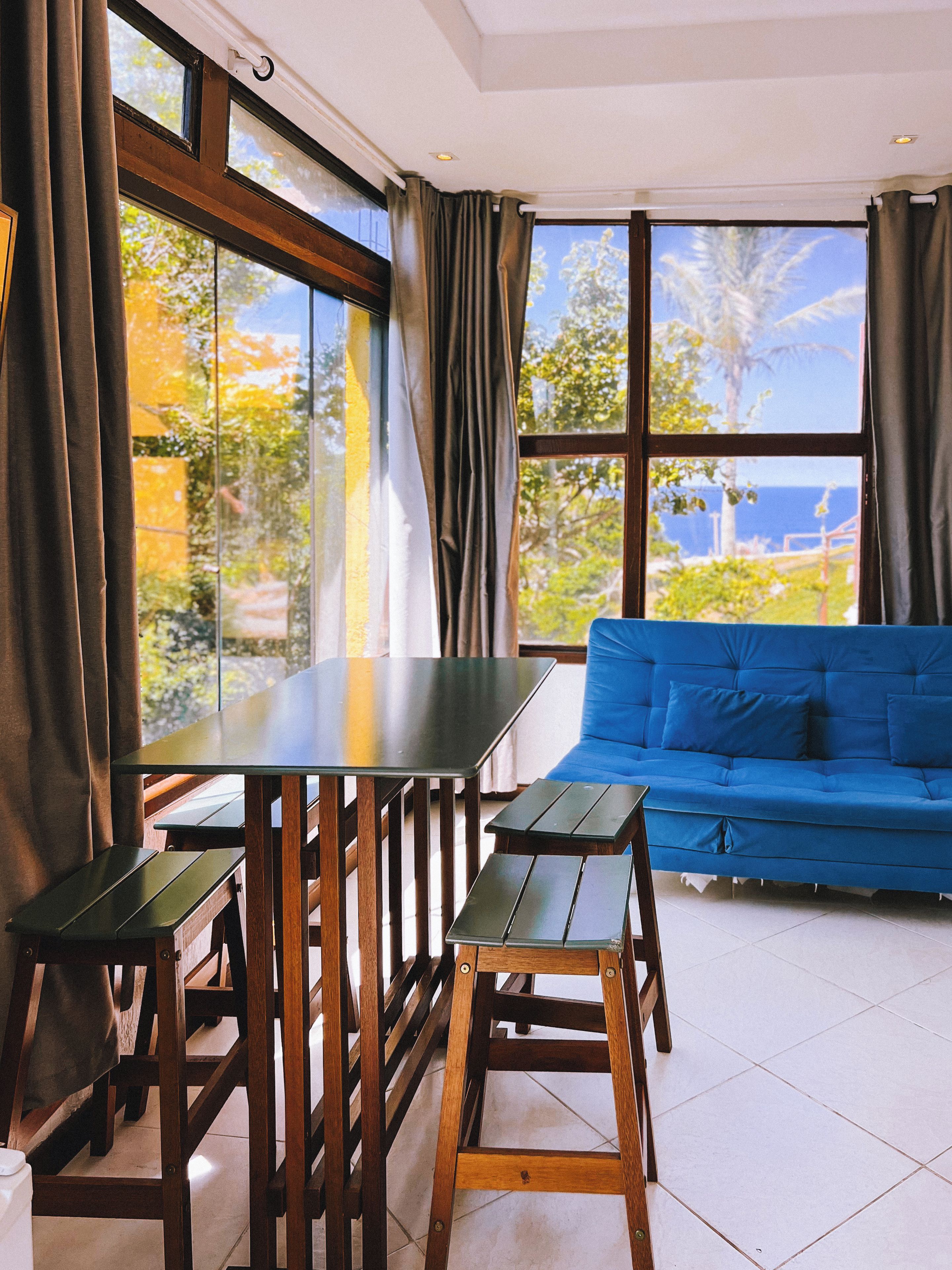 Una habitación con una mesa de madera y bancos, un sofá azul y ventanas grandes con cortinas.