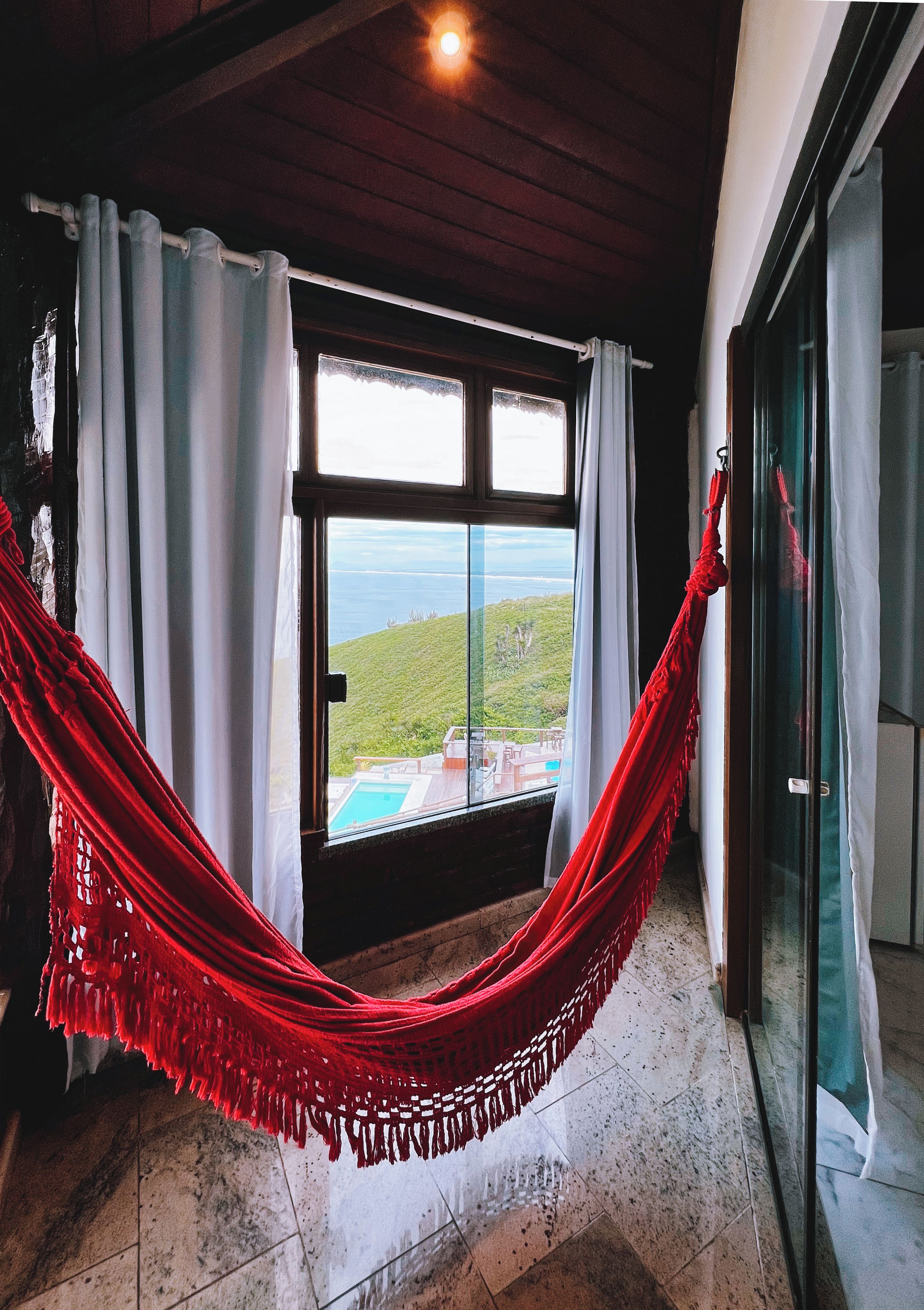 Ein Zimmer mit großem Fenster, einer roten Hängematte und Blick auf eine grüne Landschaft.