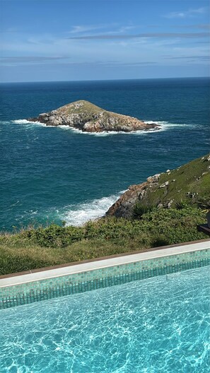 Una piscina con agua cristalina azul, rodeada de vegetación y con vistas a un litoral rocoso y al océano al fondo.